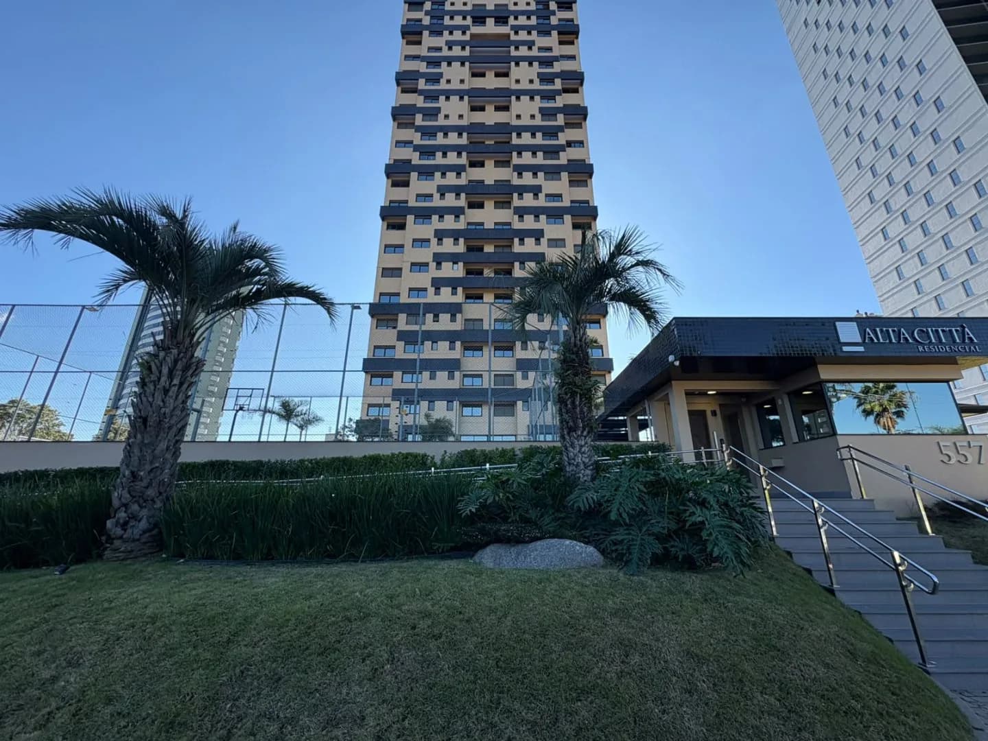 Entrada do prédio com escada de acesso, jardim e palmeiras.