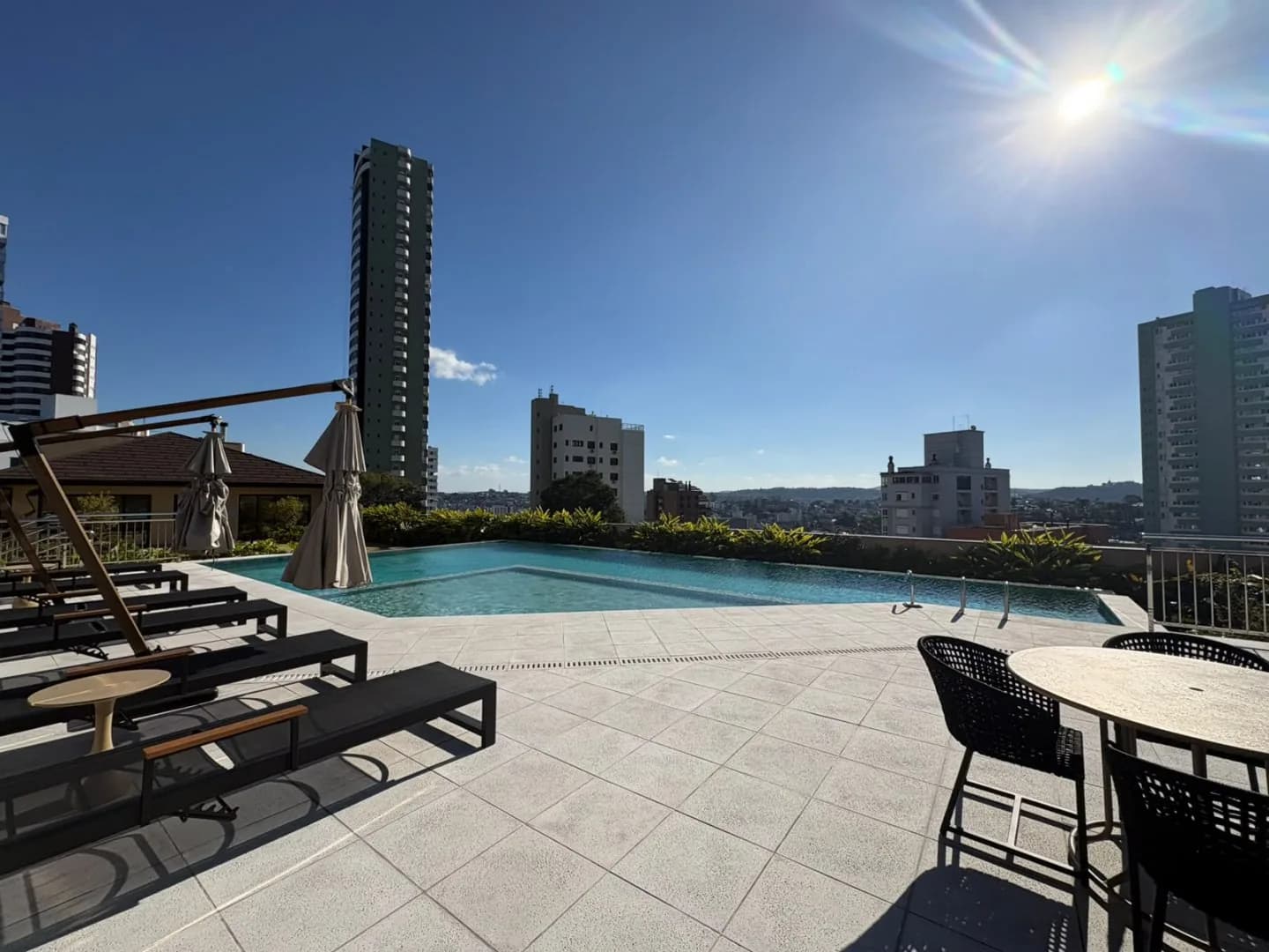 Piscina com deck, espreguiçadeiras e mesa, com vista para a cidade.