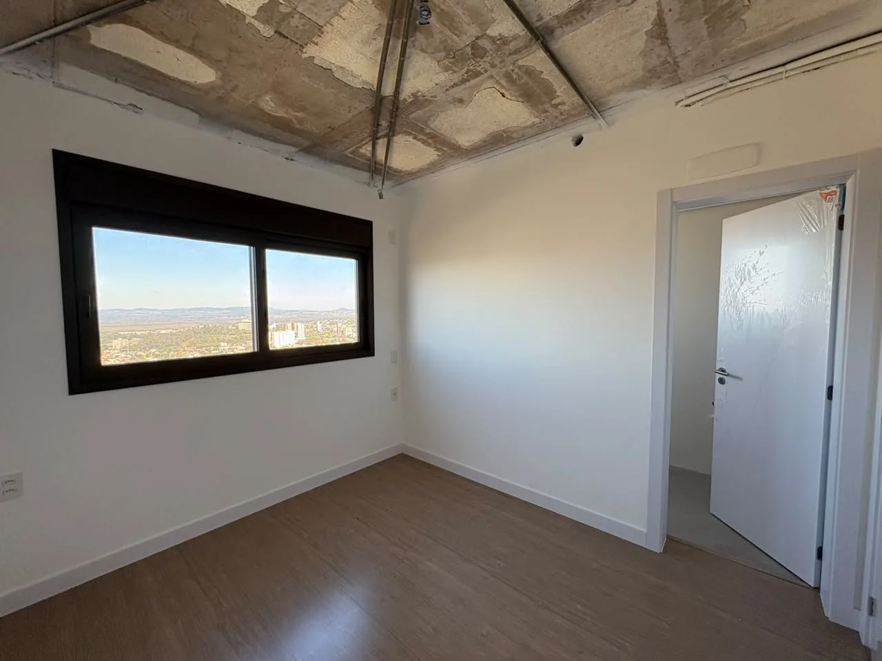 Dormitório com janela ampla, piso vinílico e porta do banheiro.