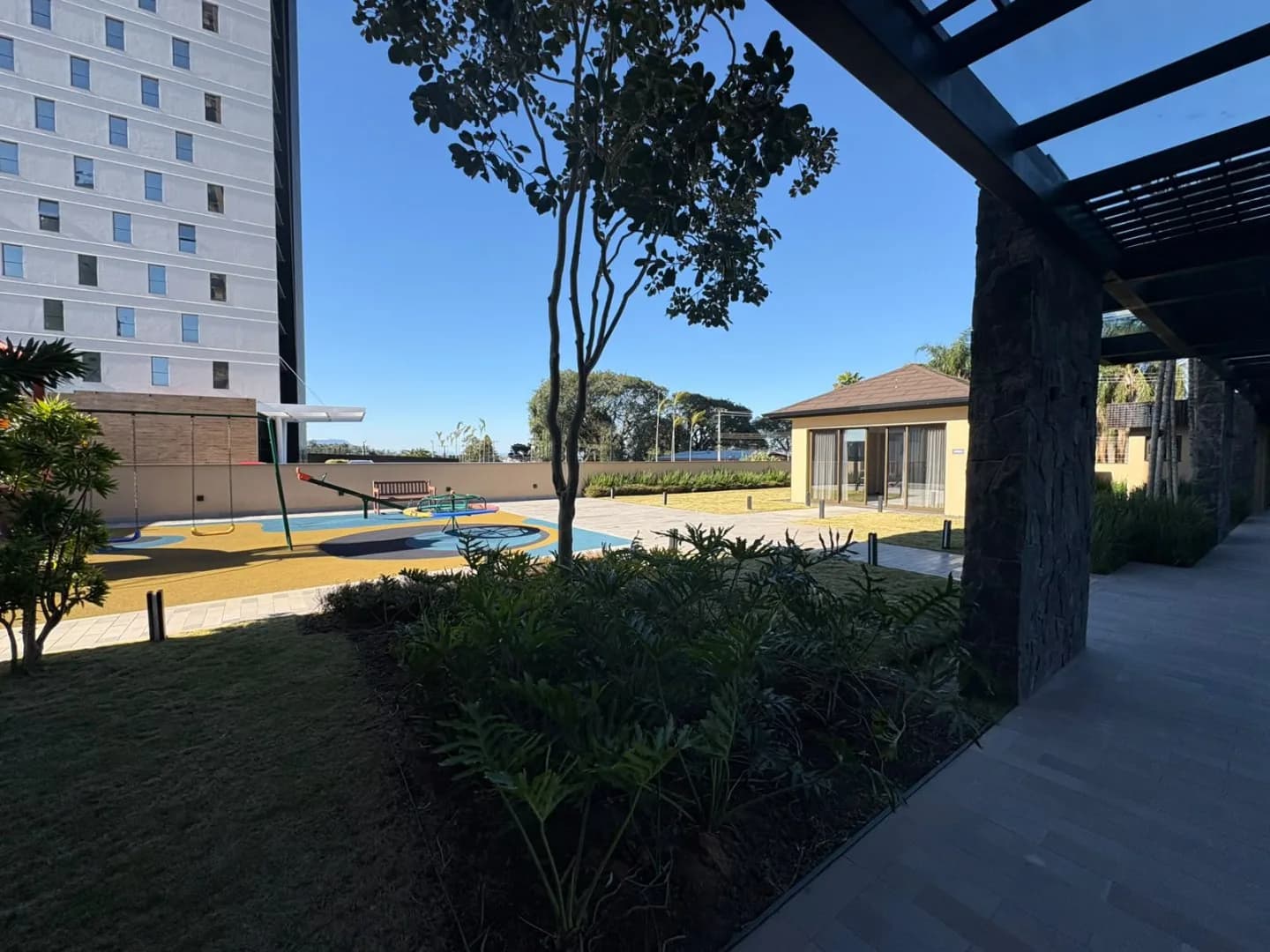 Jardim do condomínio com playground e salão de festas ao fundo.