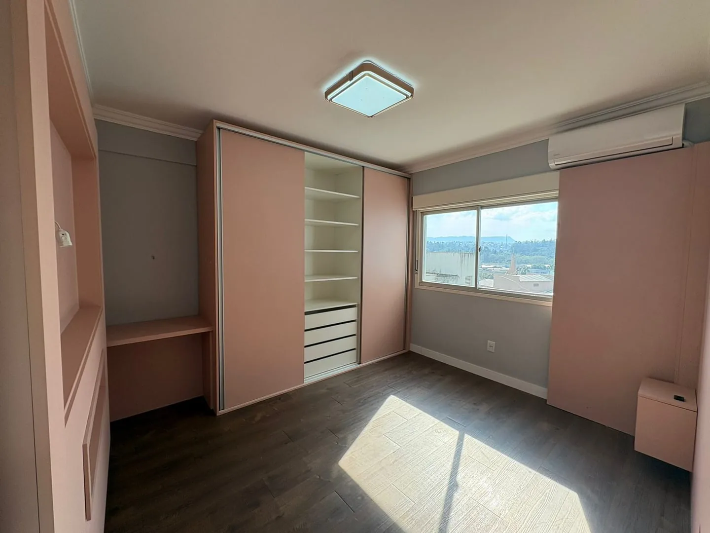 Dormitório com armário rosa embutido, bancada e piso laminado escuro.