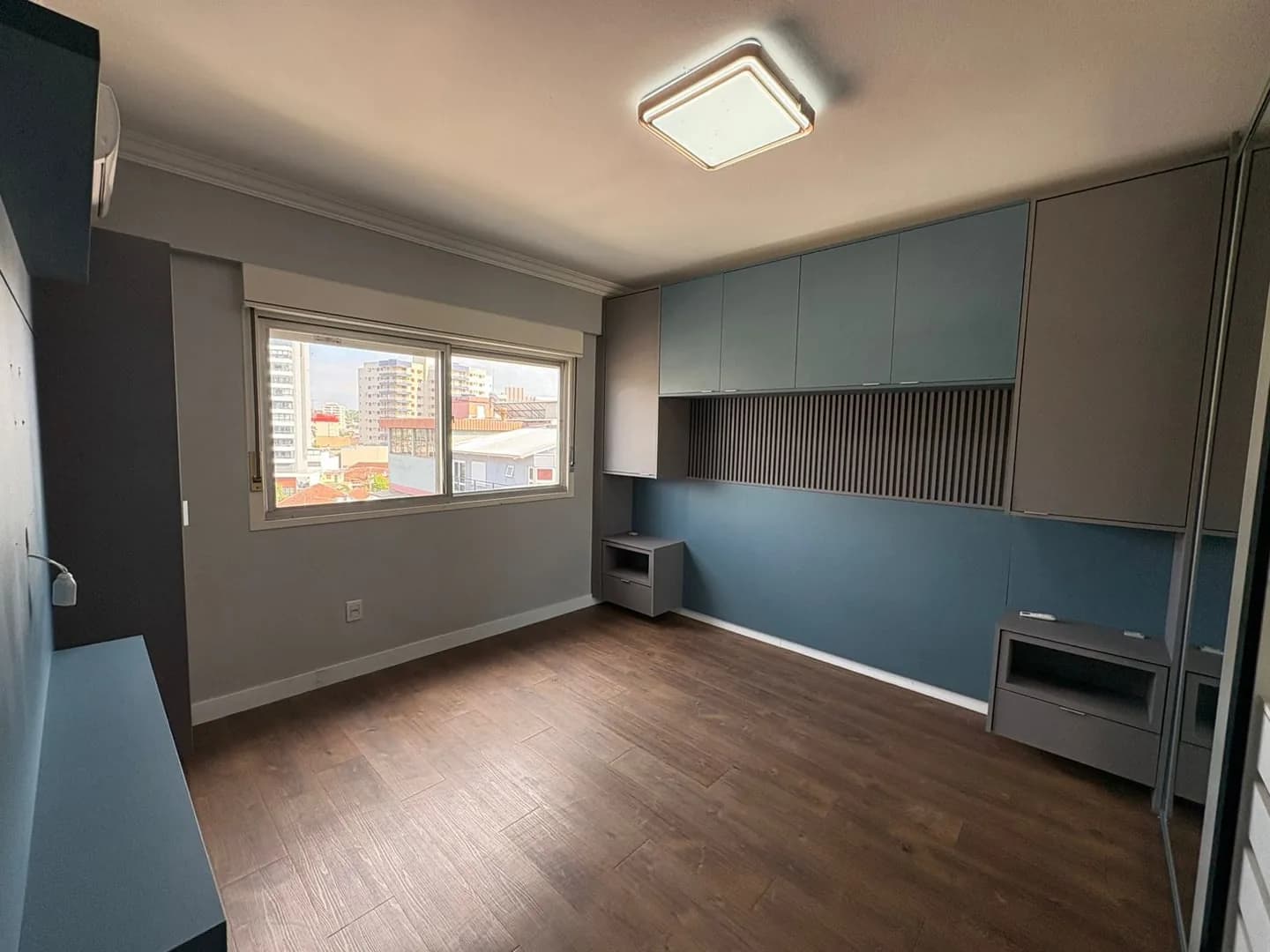 Dormitório com armários azuis e cinza, janela ampla e piso laminado.