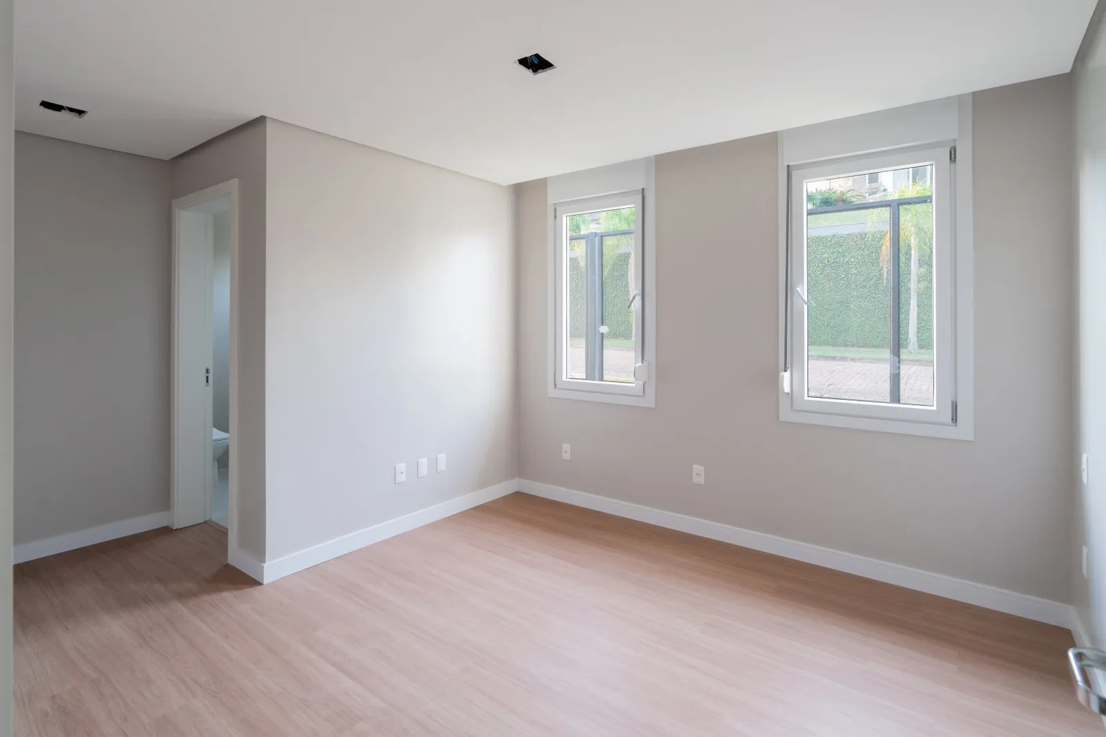 Quarto com duas janelas, piso laminado e banheiro ao lado.