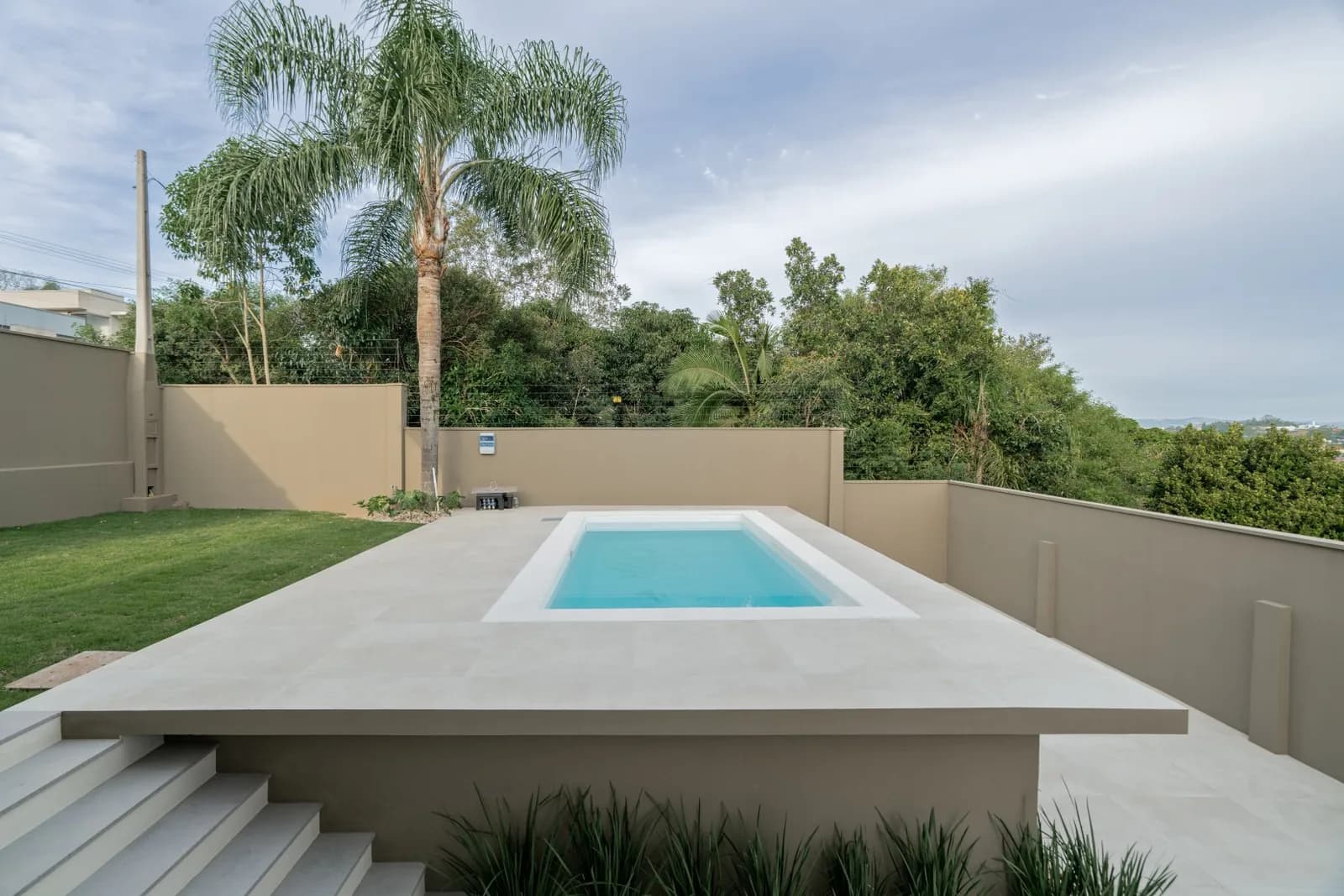 Piscina com deck de porcelanato, gramado e palmeira ao lado.