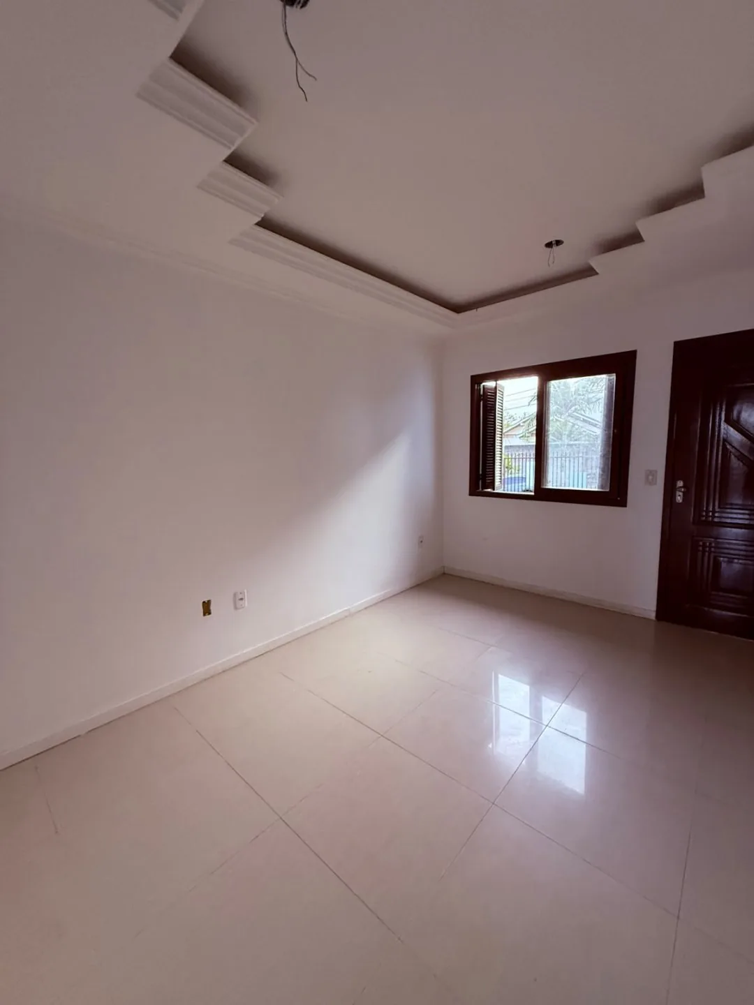 Sala vazia com piso claro, janela e porta de entrada em madeira.