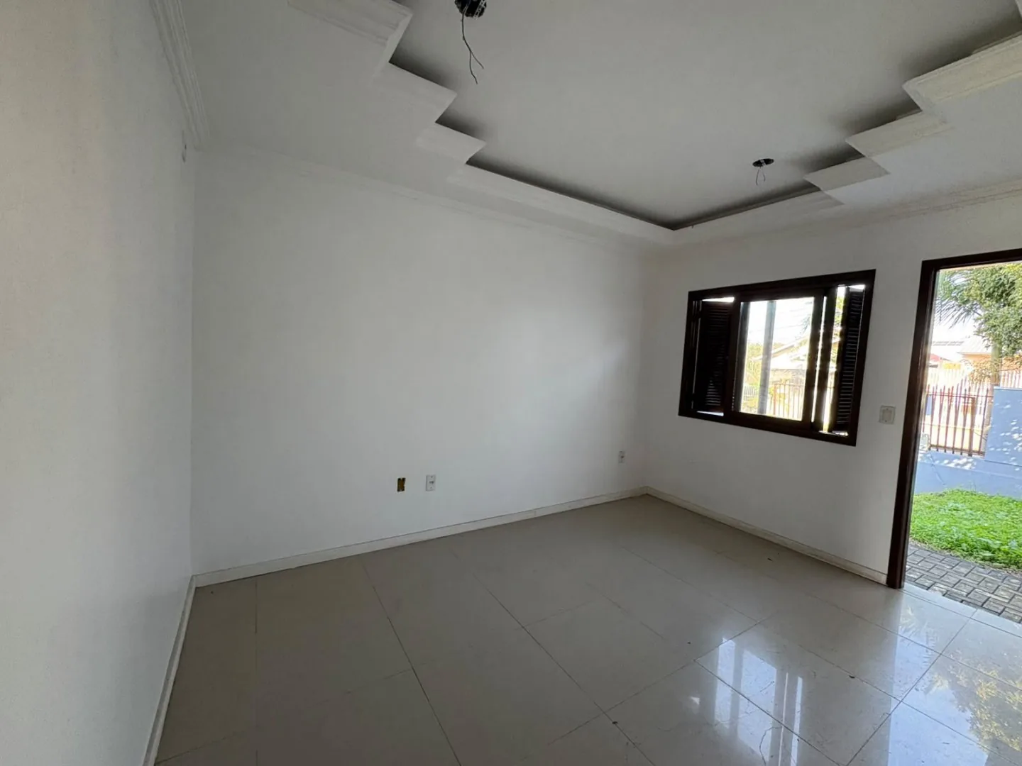 Sala vazia com piso claro, janela e porta que dá para o pátio da frente.