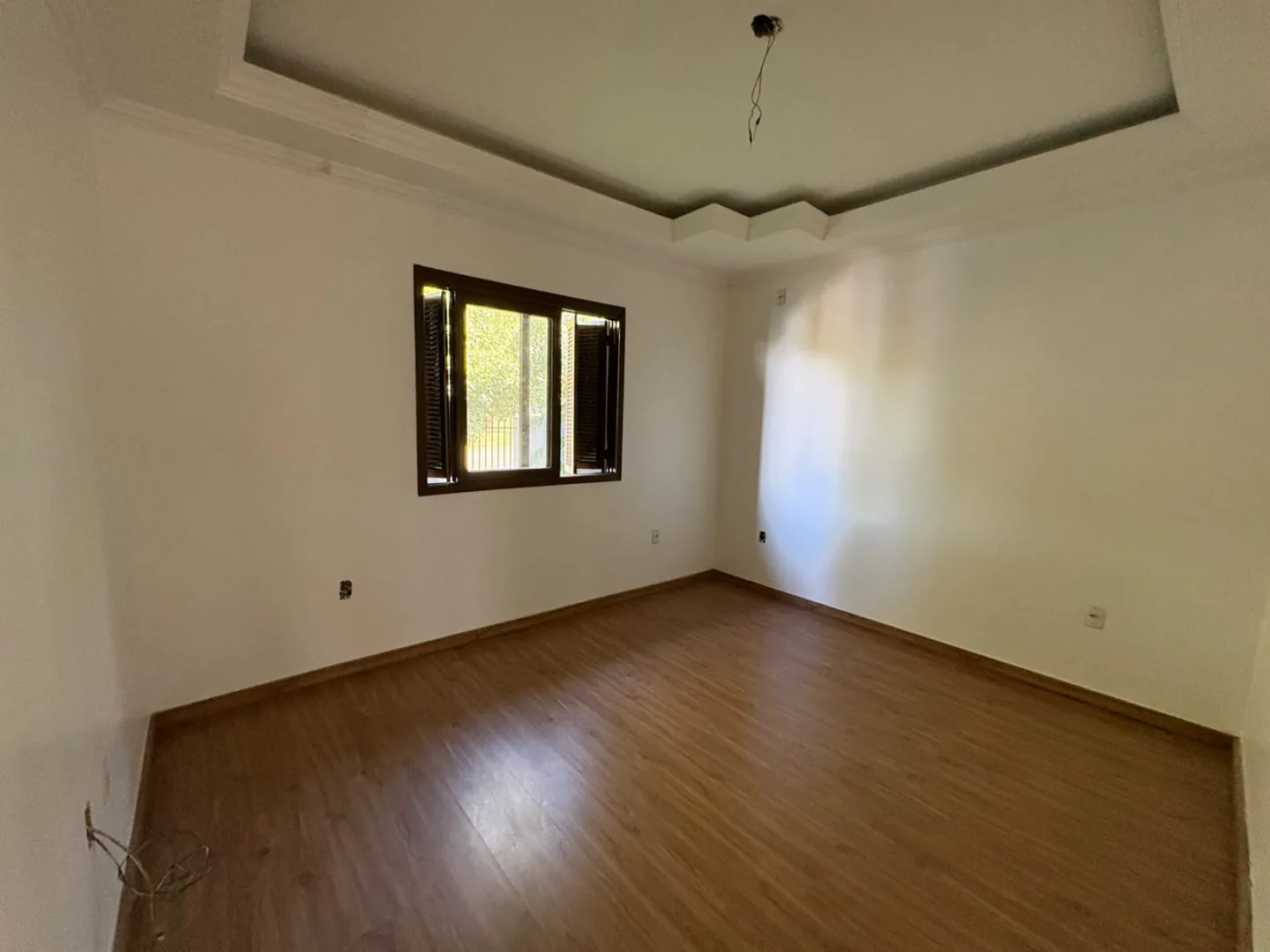 Dormitório vazio com piso laminado, sanca de gesso e janela de madeira.