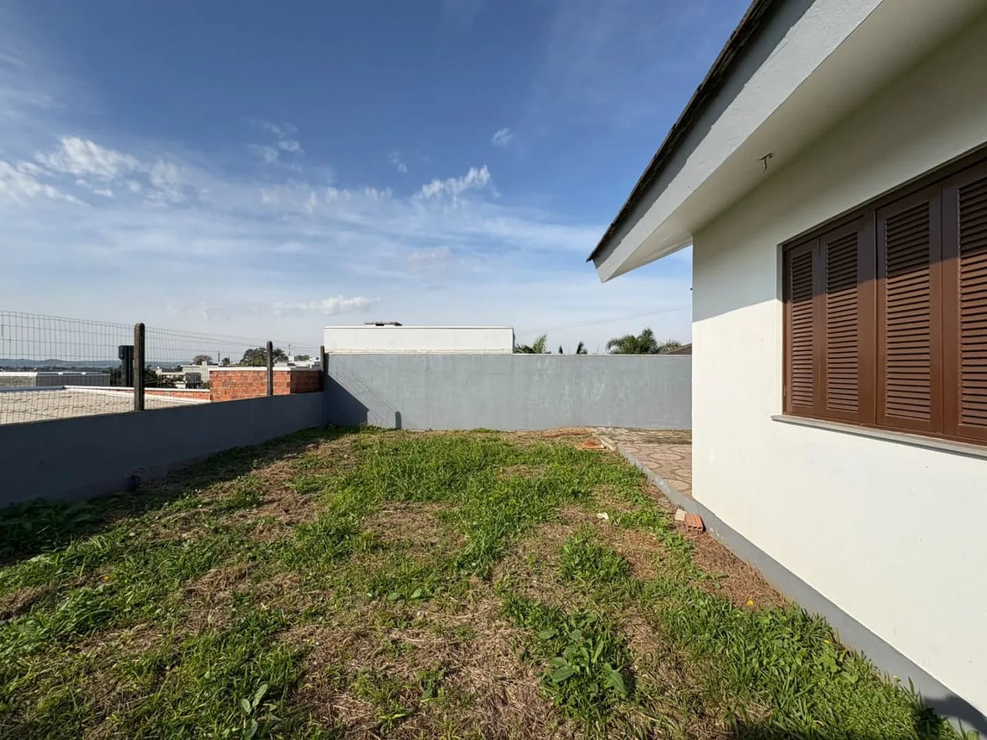Pátio lateral gramado, com muro, tela e janela de madeira da casa.