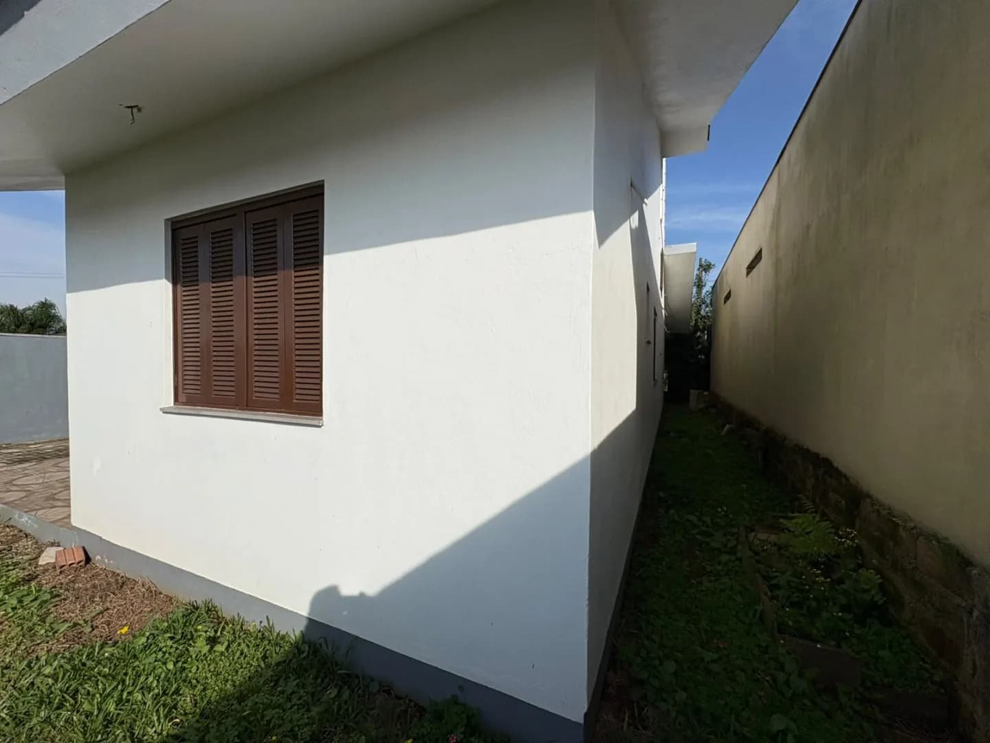 Lateral da casa com janela de madeira e faixa de grama junto ao muro.