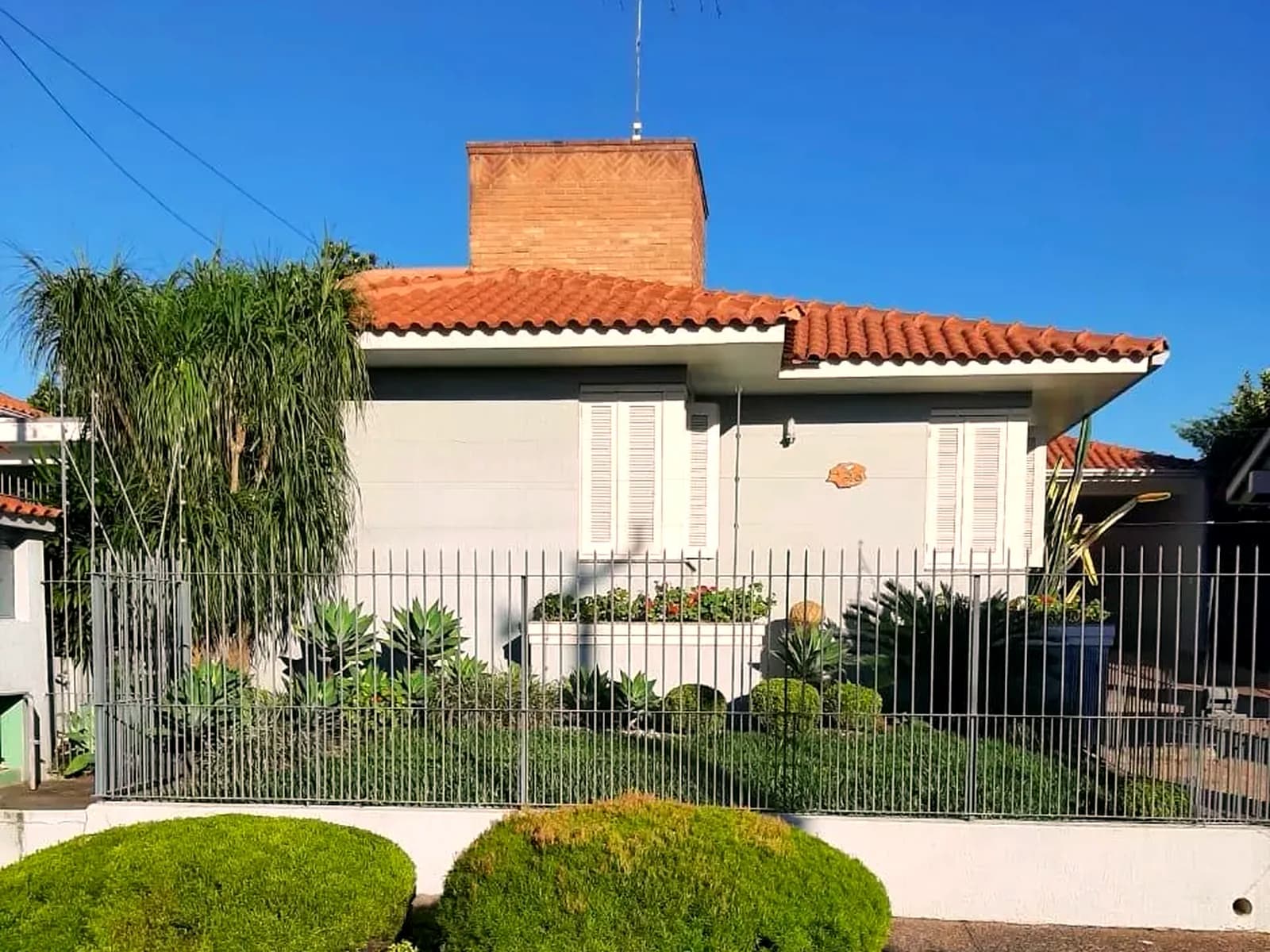 Fachada branca com telhado de barro, grades e o jardim aparado na frente.