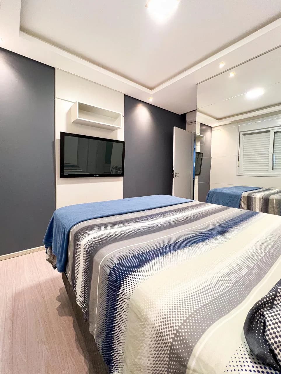 Dormitório principal, com cama de casal, TV de parede e roupa de cama listrada azul.