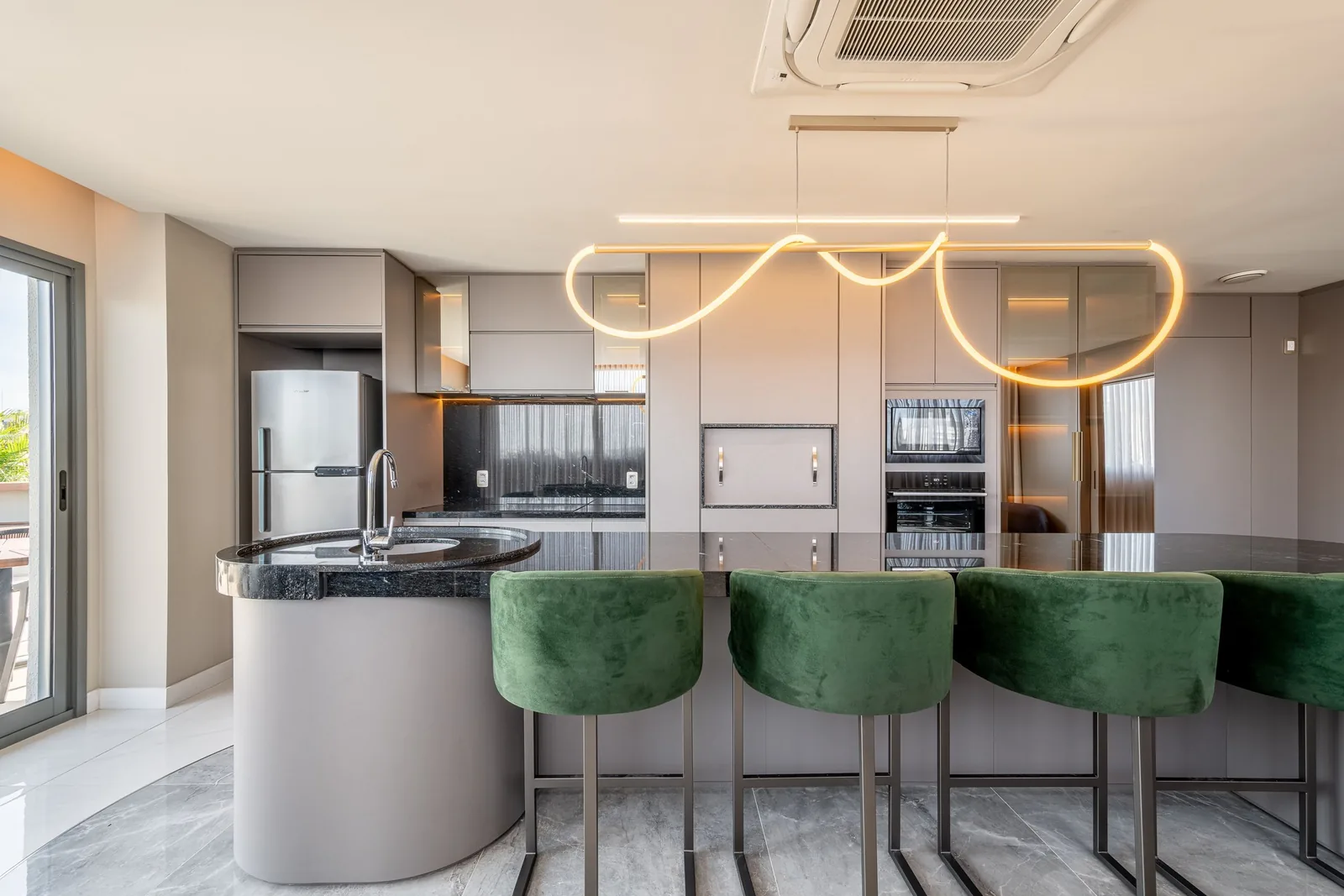 Espaço gourmet com bancada, banquetas de veludo verde e cozinha equipada.