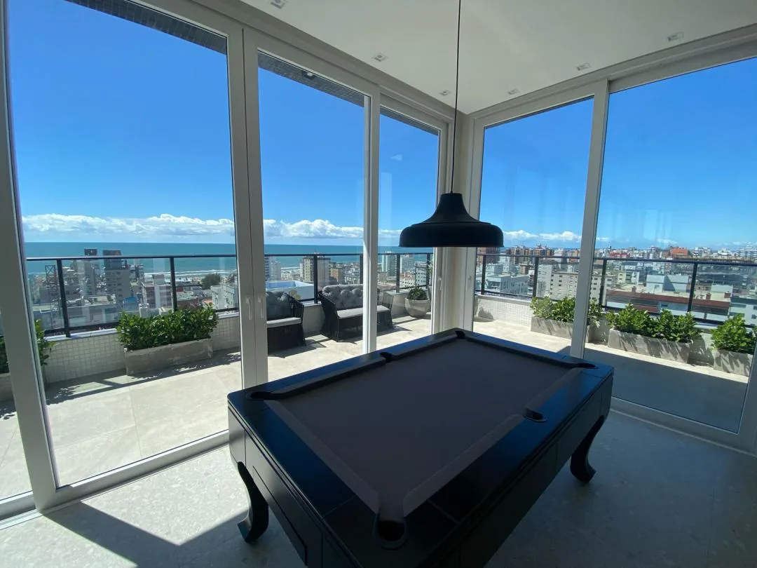 Salão com mesa de sinuca e janelas de vidro com vista para o mar.