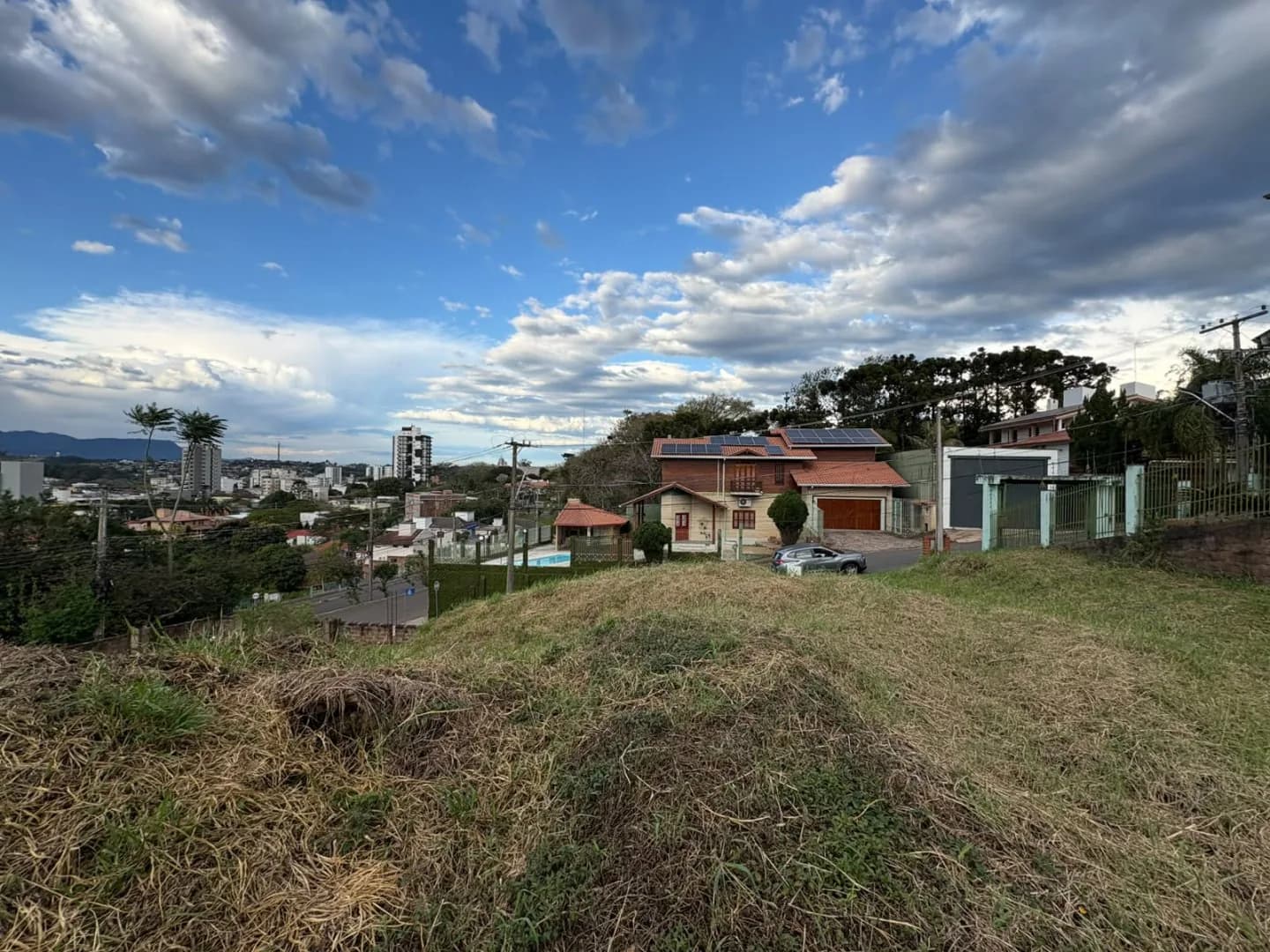 Terreno no alto, casa vizinha de telhado laranja e cidade ao fundo.