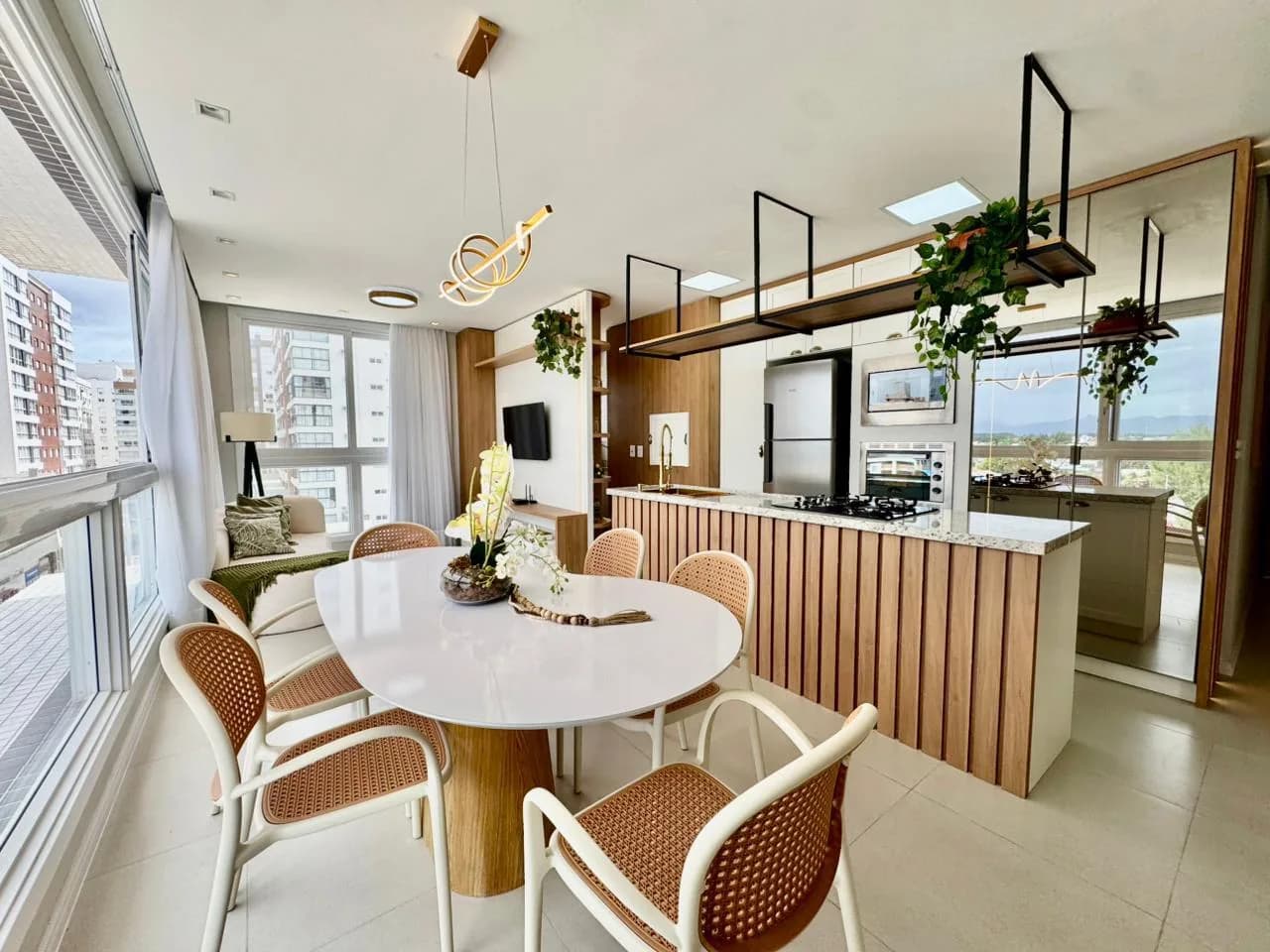 Living integrado com mesa de jantar branca, cadeiras de palhinha, ilha da cozinha com ripado de madeira e estar com TV ao fundo