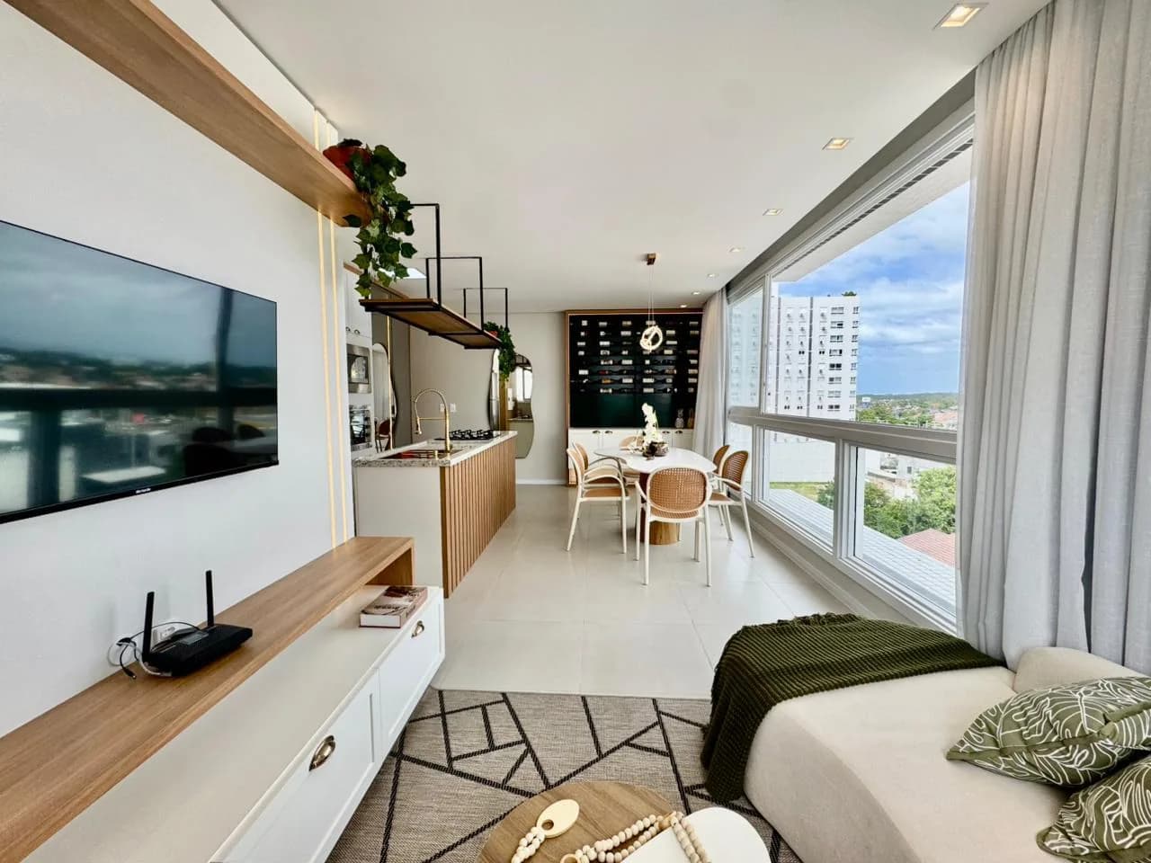 Sala de TV com rack em madeira e sofá claro, com a mesa de jantar, a cozinha e a adega ao fundo