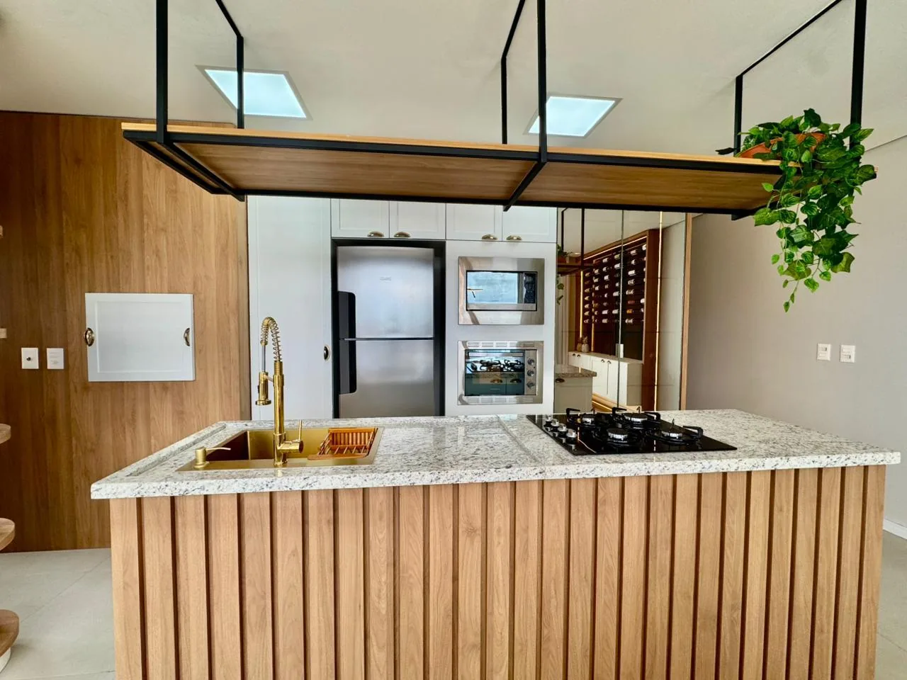 Ilha com ripado de madeira, bancada de granito e cooktop a gás, com geladeira inox, micro-ondas e forno embutidos ao fundo