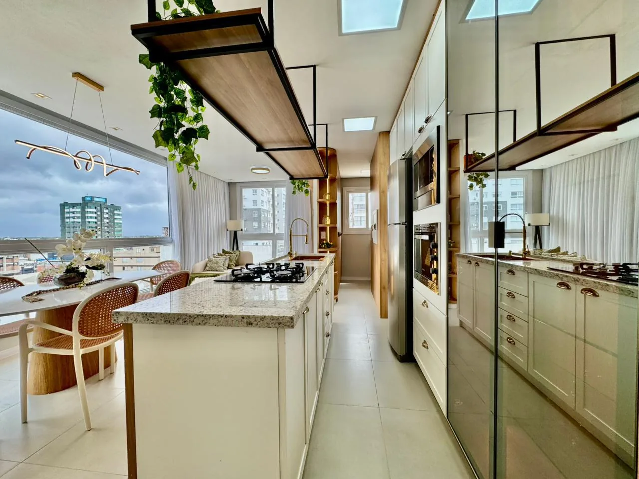 Cozinha com armários brancos, ilha com cooktop e bancada de granito, prateleira suspensa com plantas e mesa de jantar junto à janela