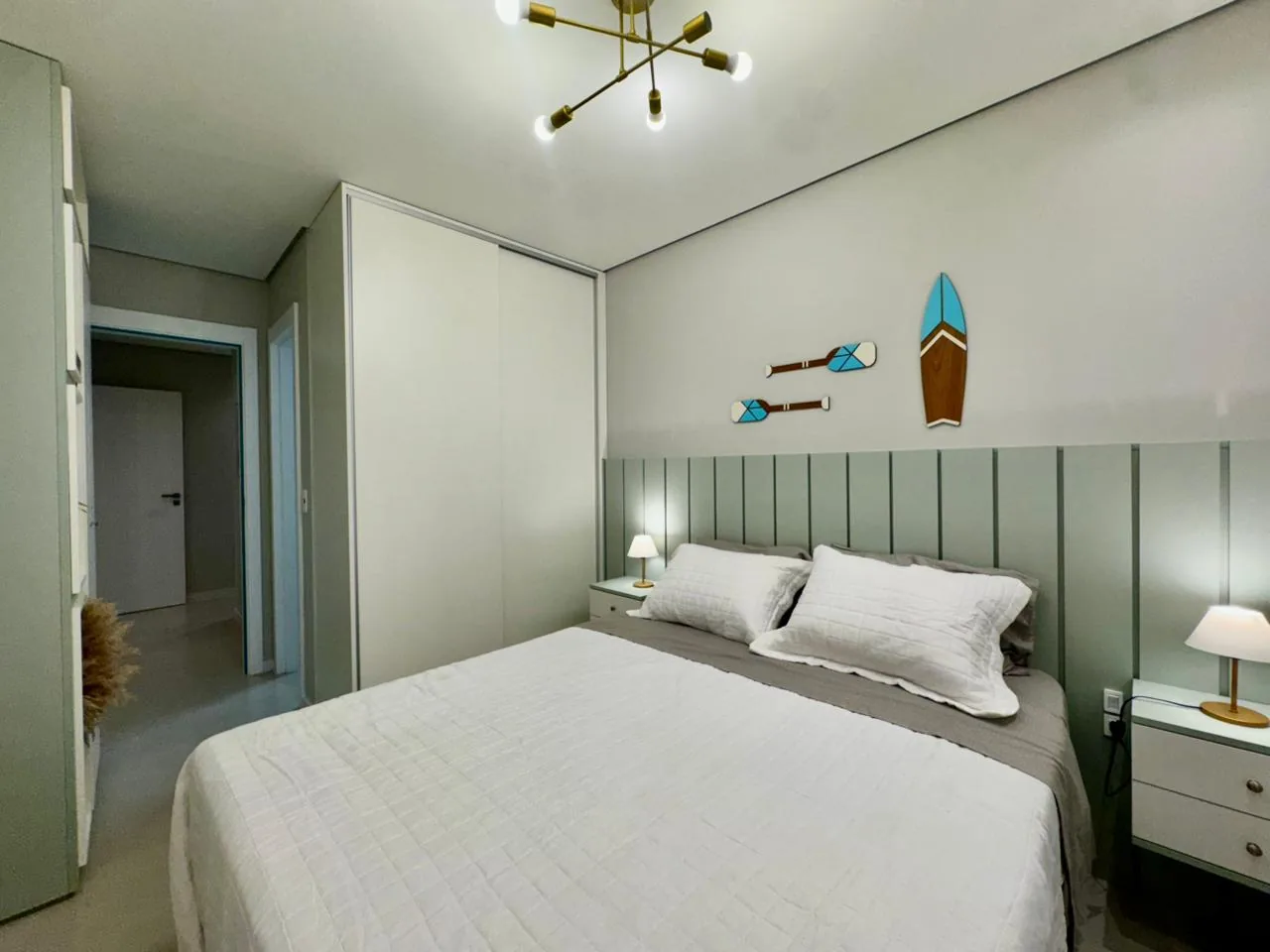 Suíte com cabeceira em painel verde, prancha de surfe e remos decorativos na parede e armário branco