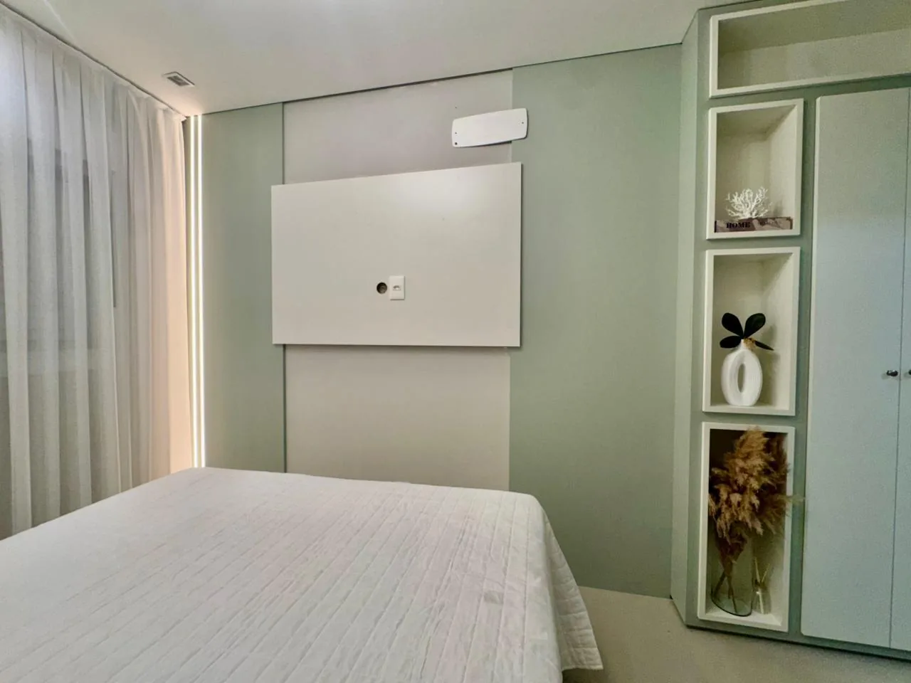 Quarto com parede verde-clara, painel branco para TV e nichos decorativos ao lado do armário