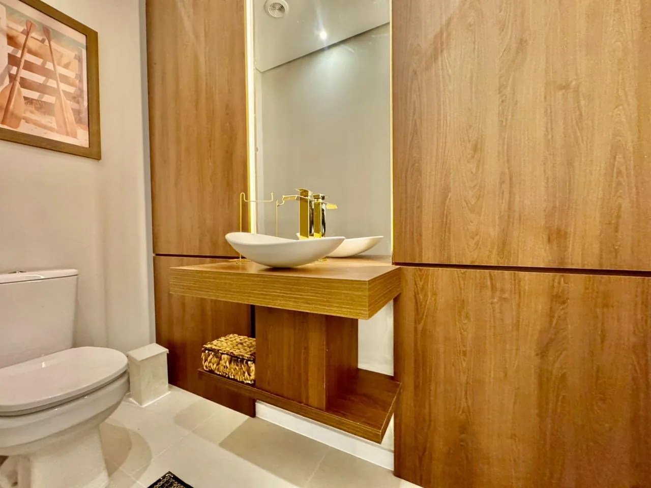 Lavabo com painéis de madeira, cuba de apoio branca, torneira dourada e espelho com luz embutida