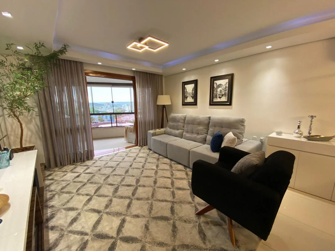 Sala de estar com sofá cinza, poltrona preta, tapete geométrico e forro rebaixado com LED; ao fundo, a porta para a sacada envidraçada com vista da cidade