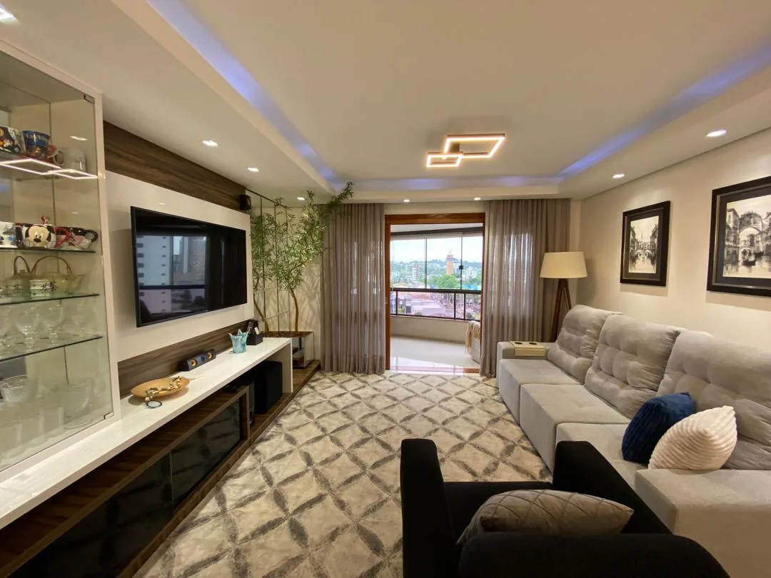 Sala com painel de TV em madeira, cristaleira de vidro iluminada e sofá cinza, com a sacada envidraçada ao fundo
