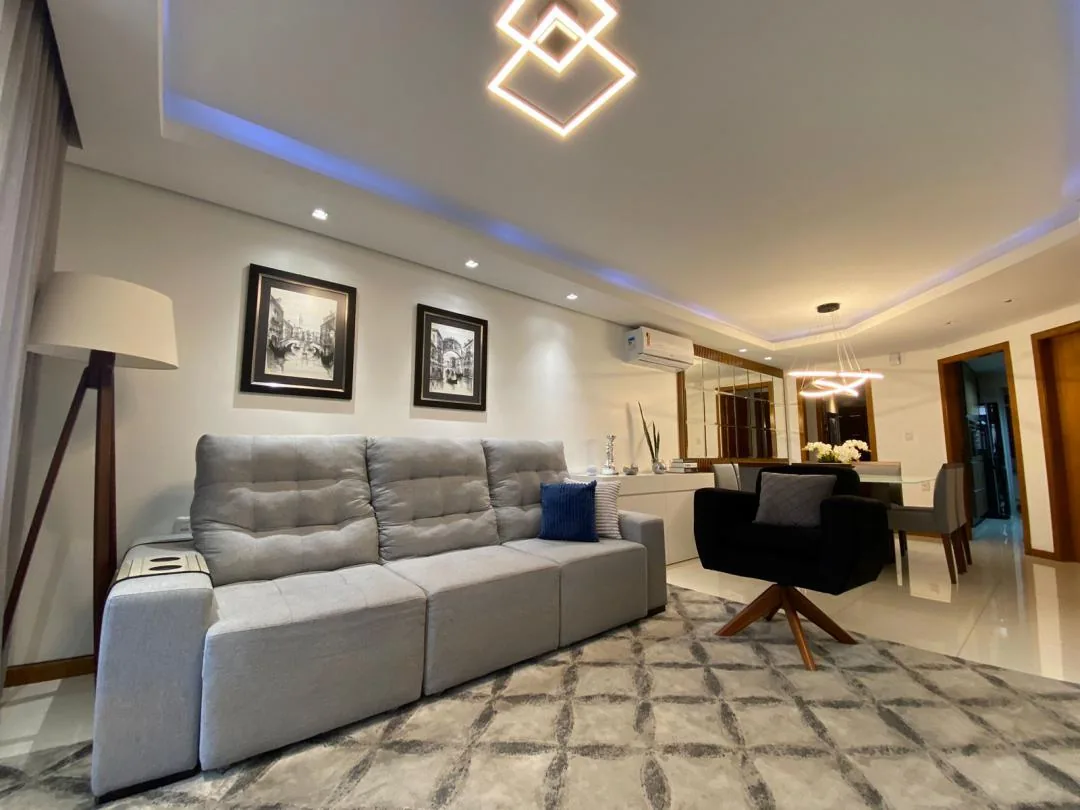Sofá cinza sob dois quadros em preto e branco, luminária de pé e luminária de LED no forro, com a mesa de jantar ao fundo