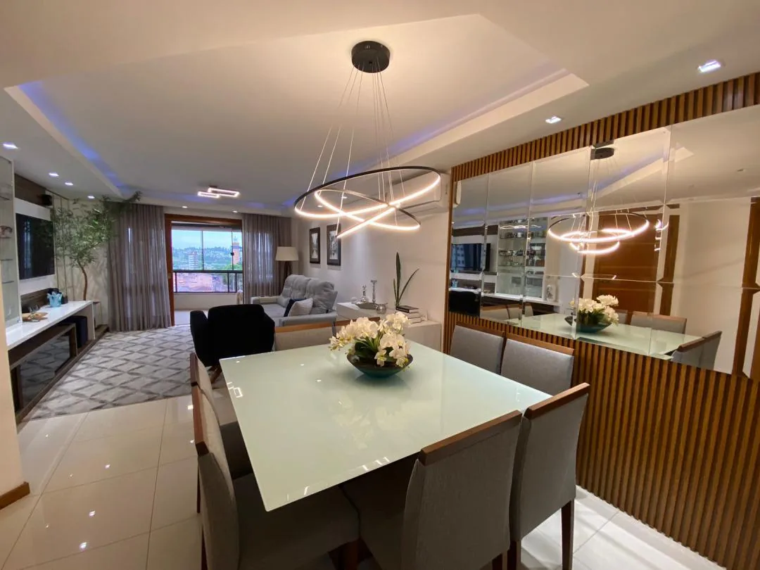 Sala de jantar com mesa de tampo de vidro, cadeiras cinza, lustre de anéis de LED e parede de espelhos com ripado de madeira, integrada ao estar