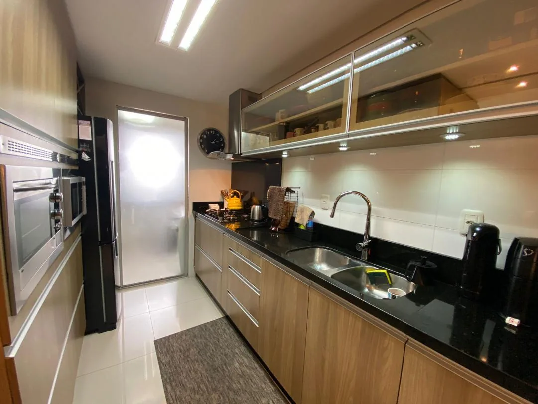 Cozinha com armários em madeira clara, bancada de granito preto com cuba de inox, forno e micro-ondas embutidos e geladeira inox