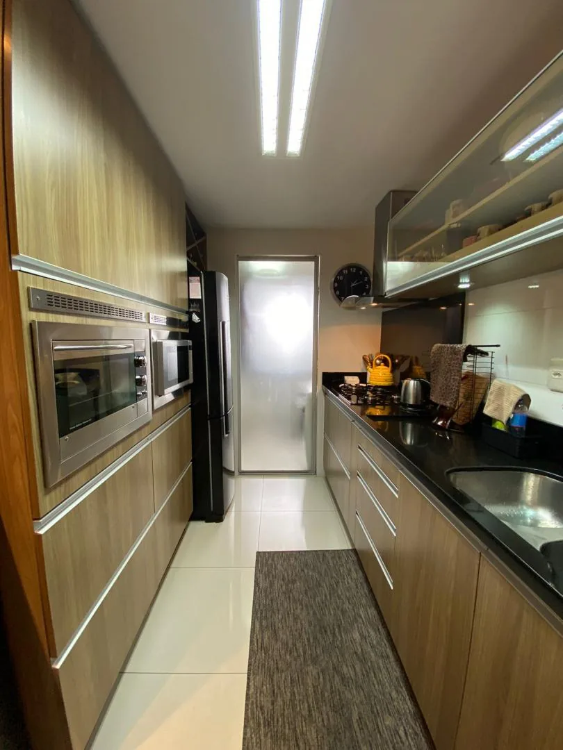 Cozinha em corredor com torre de forno e micro-ondas embutidos, bancada preta com cooktop e porta de vidro jateado ao fundo