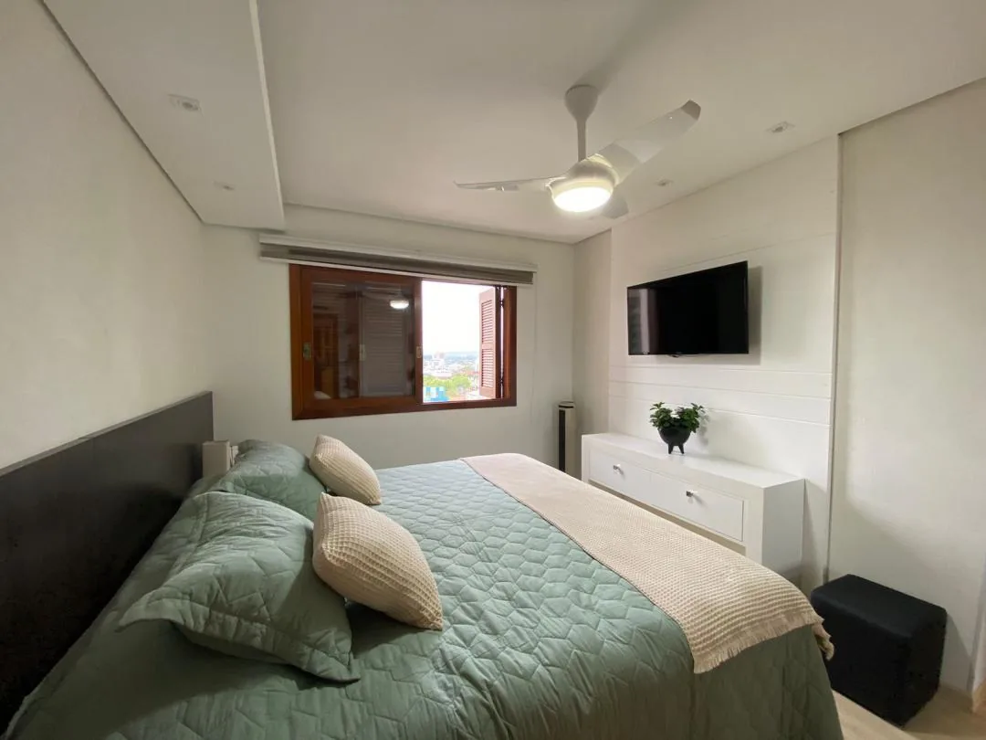 Quarto com cama de casal, colcha verde, cabeceira escura, ventilador de teto, TV na parede e janela com vista da cidade