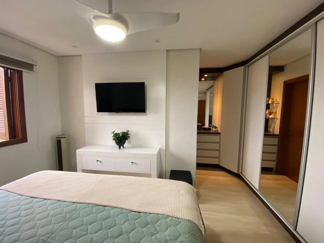Quarto com TV sobre cômoda branca, piso vinílico claro e armário de portas espelhadas junto à passagem para os gaveteiros