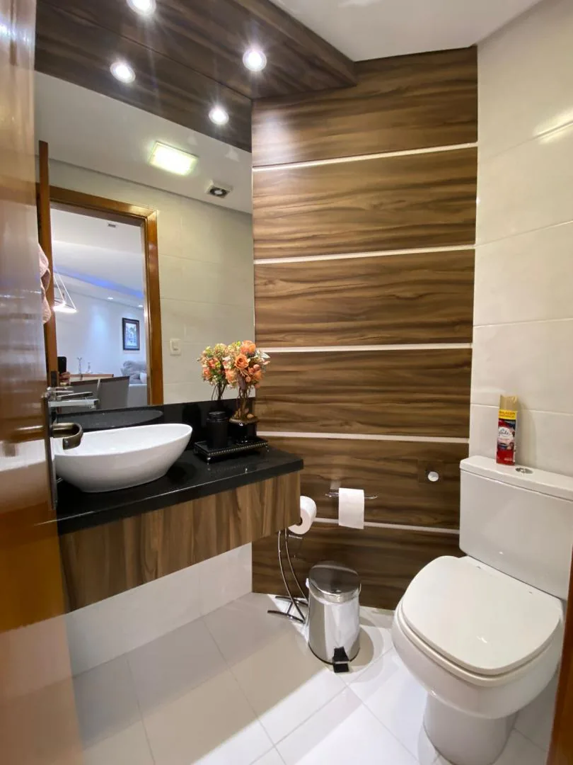 Lavabo com parede revestida em madeira com frisos, cuba de apoio branca sobre bancada preta e espelho