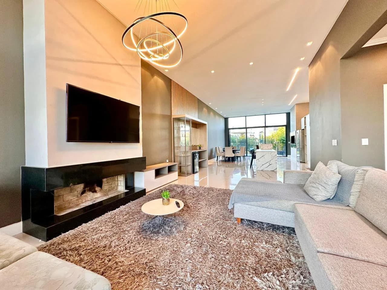 Sala de estar com lareira em moldura preta, TV na parede, lustre de anéis de LED e sofá cinza em L sobre tapete felpudo