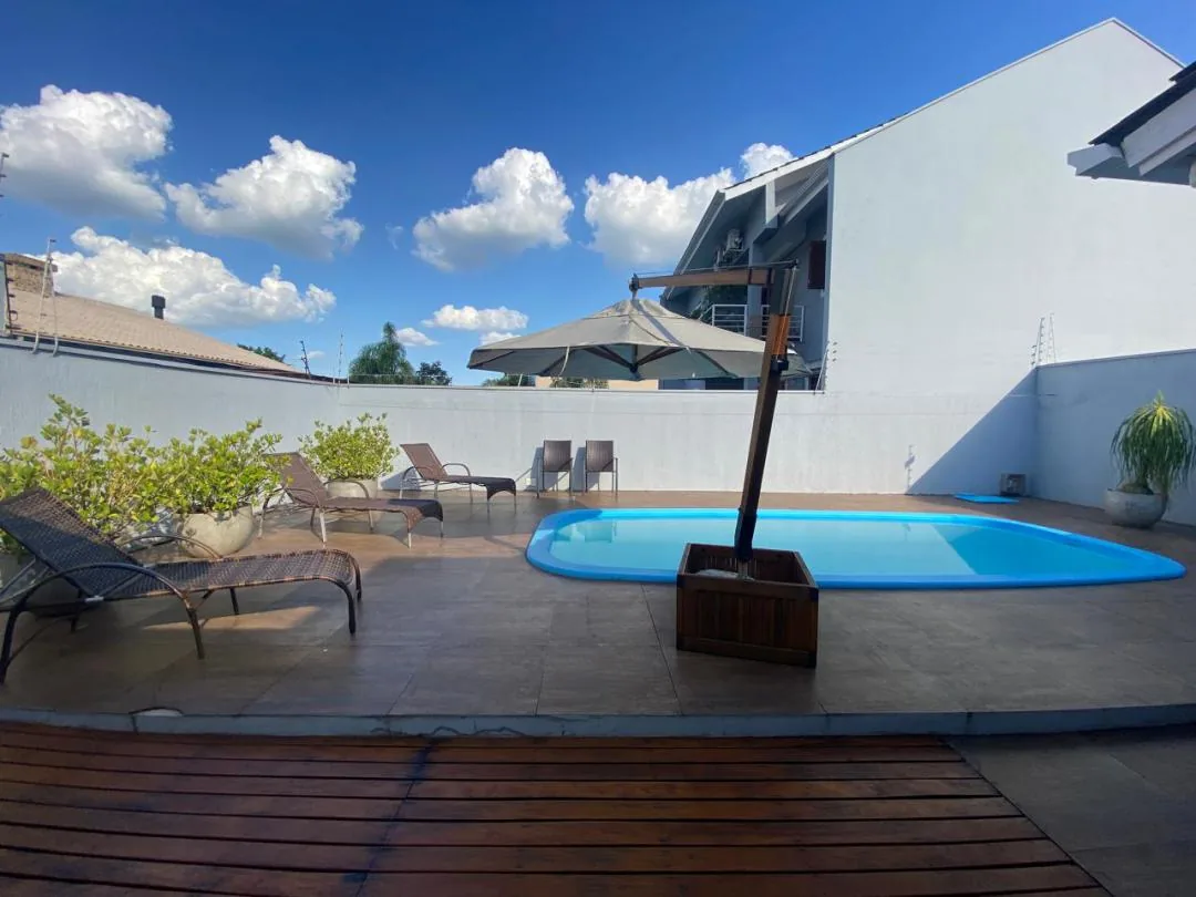 Deck de madeira em primeiro plano e piscina ao fundo, com espreguiçadeiras, vasos e ombrelone junto ao muro