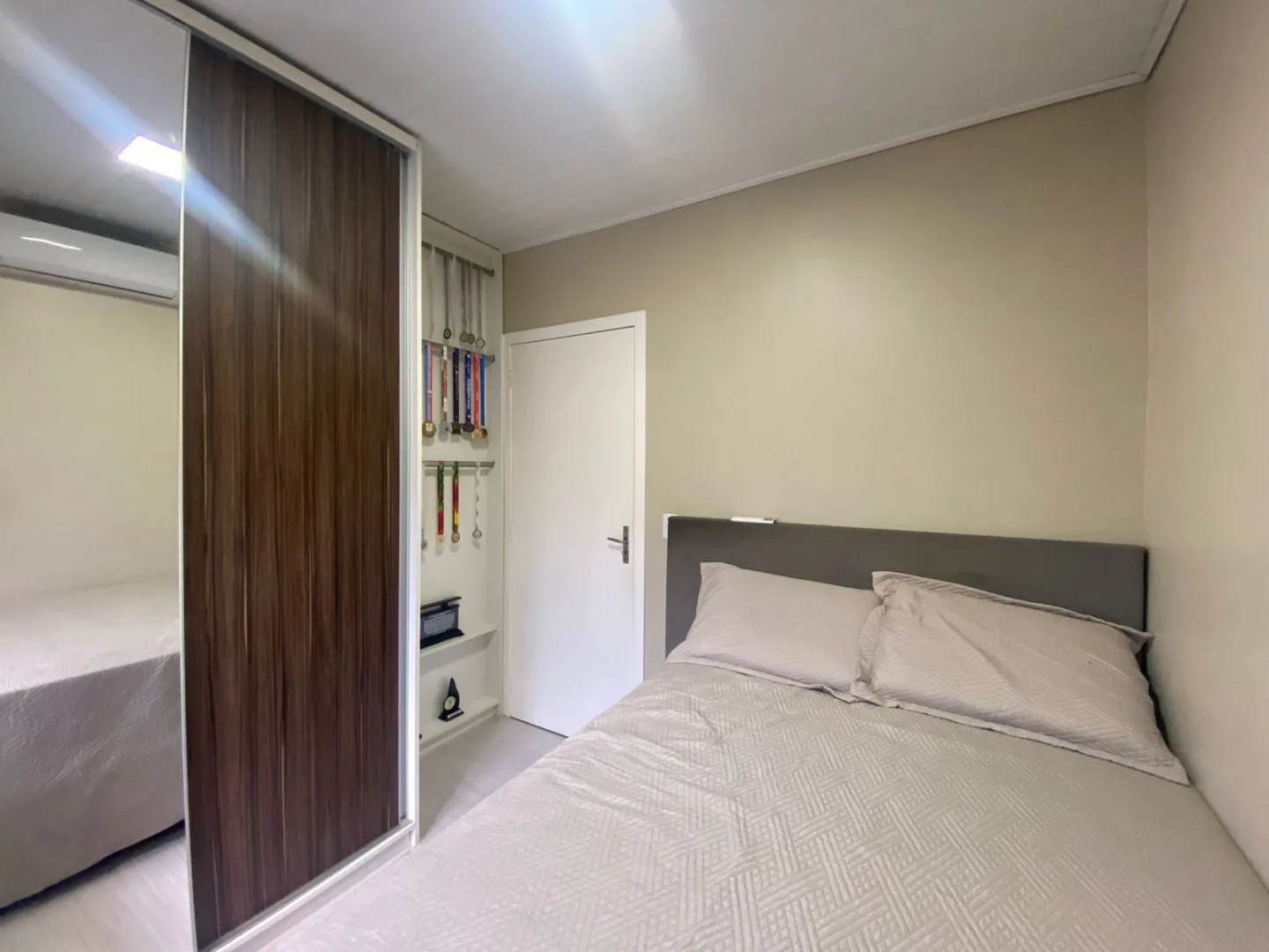 Segundo dormitório com cama de casal, cabeceira estofada cinza, guarda-roupa de porta amadeirada e nicho com prateleiras