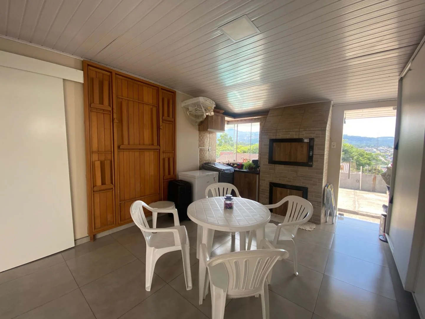 Ambiente coberto com churrasqueira de alvenaria, mesa com cadeiras brancas e porta aberta para o pátio com vista dos morros