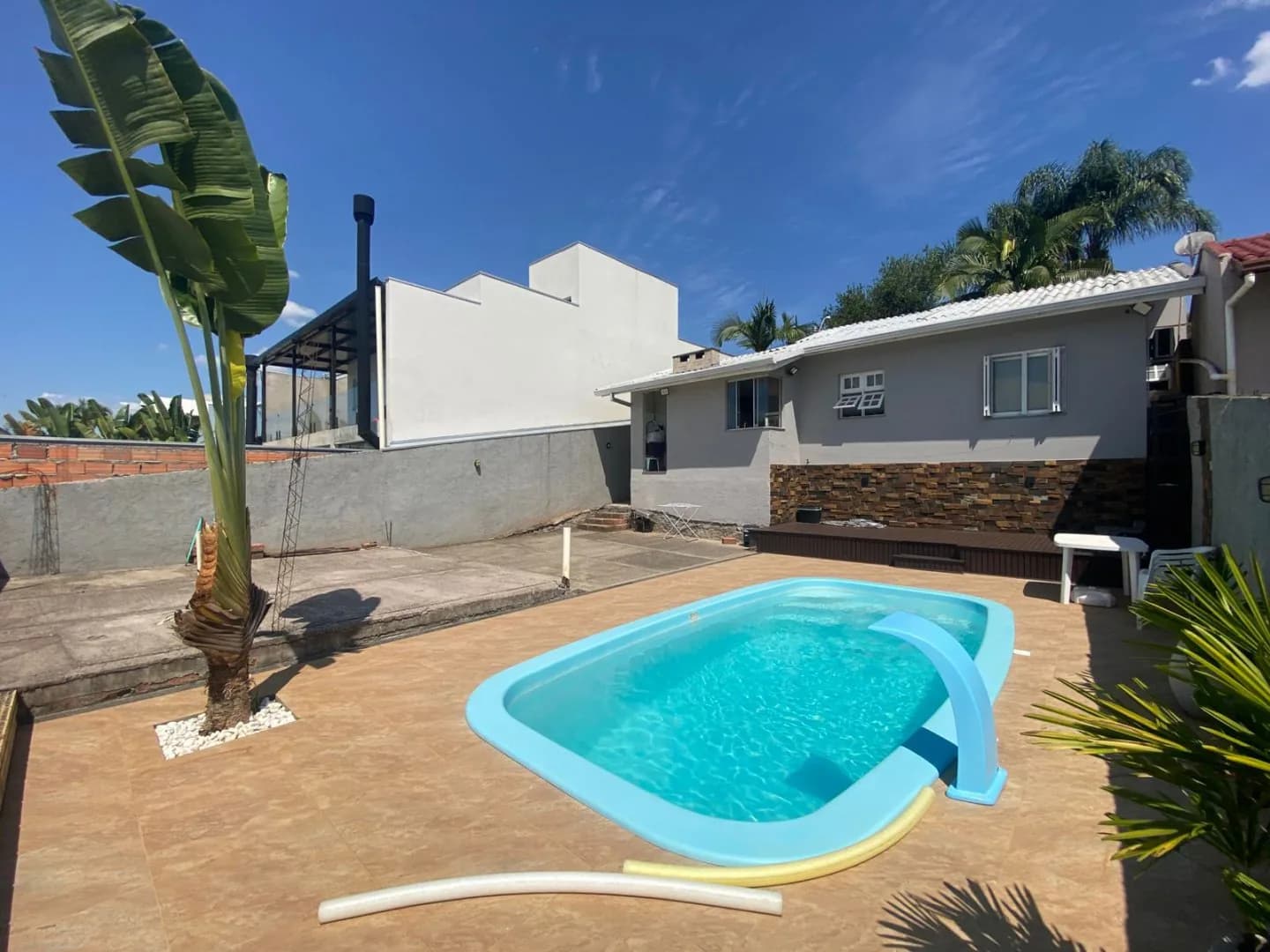 Piscina de fibra com cascata, piso cerâmico ao redor, deck de madeira e os fundos da casa com parede revestida de pedra