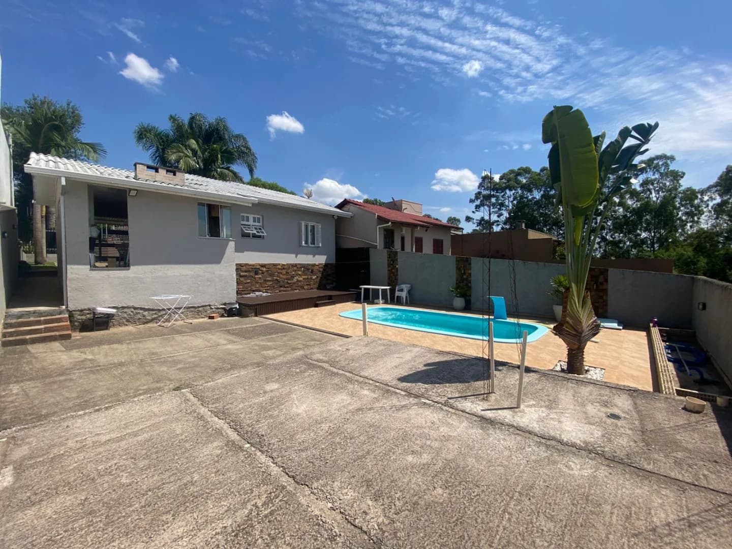Pátio dos fundos cimentado com piscina, deck de madeira e a casa cinza à esquerda