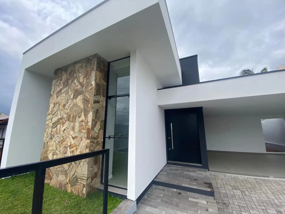 Entrada com porta preta ao lado do pilar de pedra, com a garagem coberta à direita
