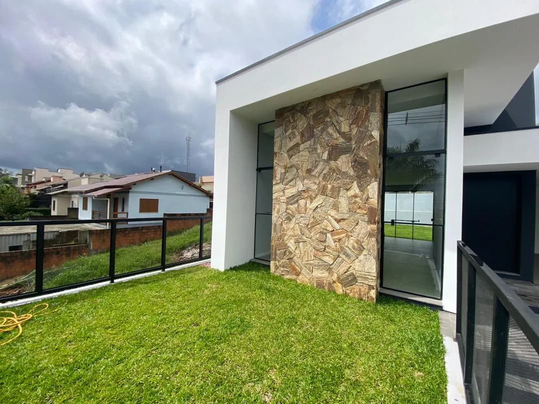 Gramado frontal elevado diante da fachada de pedra e vidro, com casas do bairro ao fundo