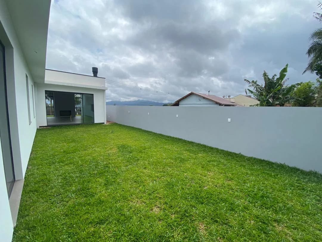Pátio gramado dos fundos com muro cinza e o living envidraçado ao fundo