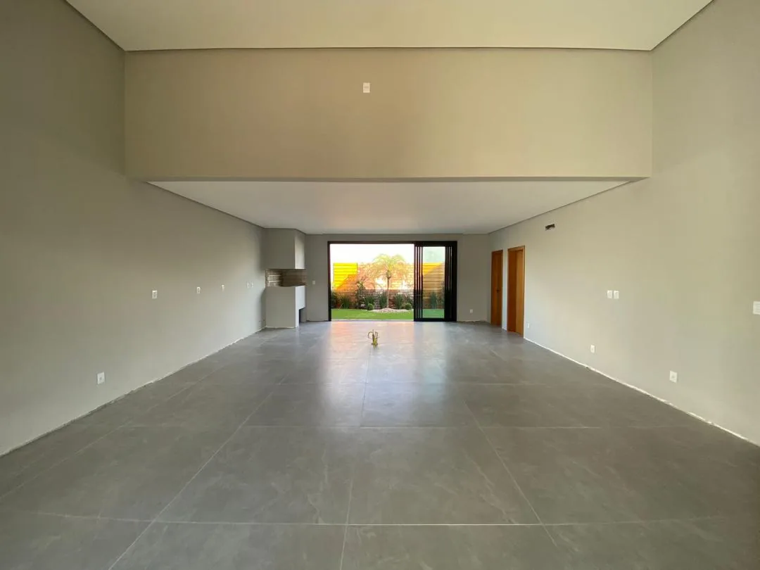 Sala ampla e vazia com porcelanato cinza em placas grandes, rebaixo de gesso e churrasqueira ao fundo, ao lado da porta de vidro que dá para o pátio