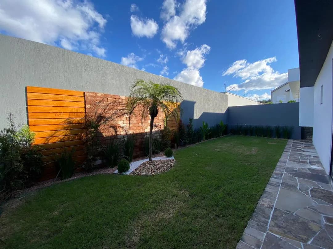 Pátio gramado com painel de madeira e muro de tijolos ao fundo, palmeira e calçada de pedra junto à casa