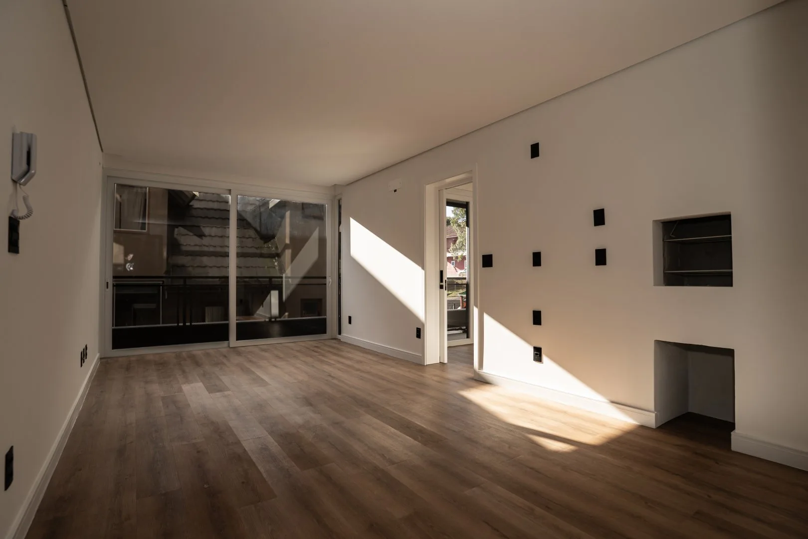 Sala com porta de vidro de correr para a sacada, nichos embutidos na parede e piso vinílico amadeirado