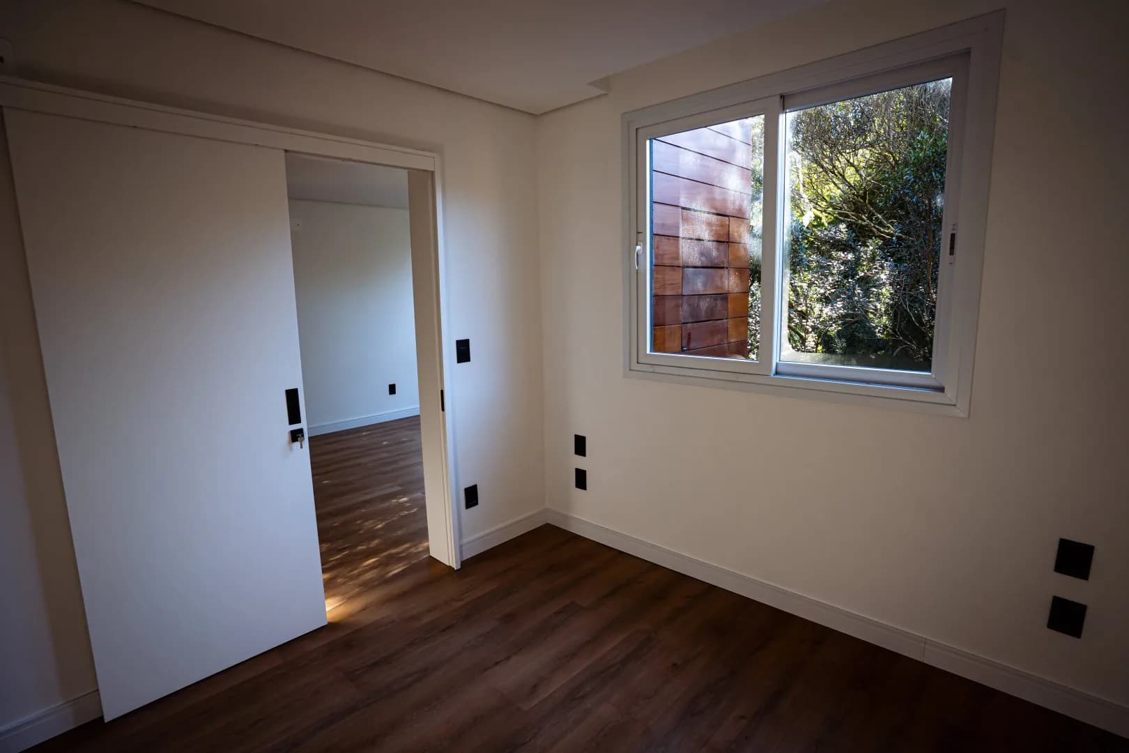 Quarto com porta de correr branca, janela de correr voltada para as árvores e piso vinílico amadeirado