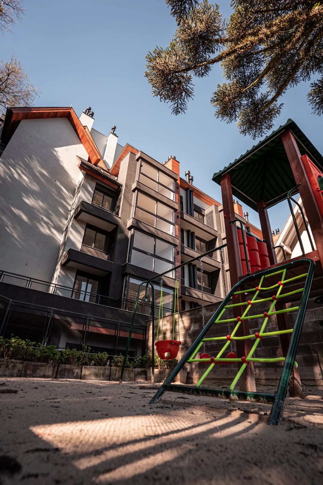 Playground externo com rede de escalada e balanço, com o prédio de fachada branca e cinza e beirais de madeira ao fundo