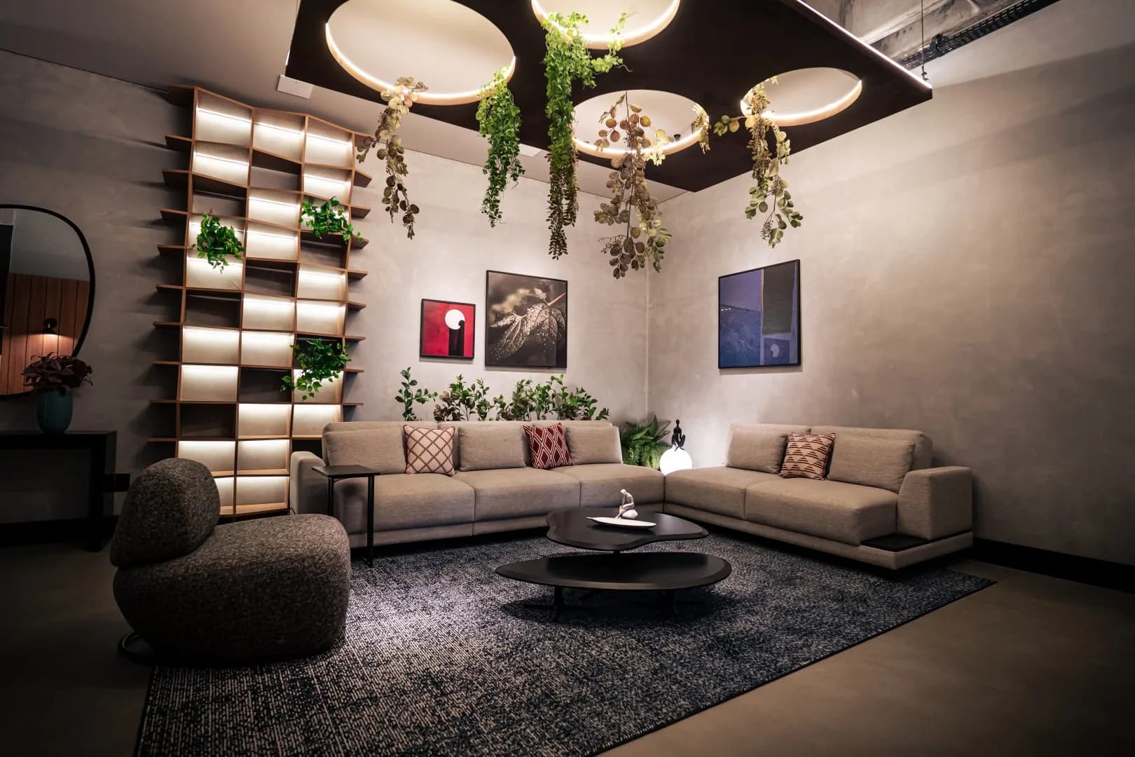 Lounge com sofás claros em L, poltrona escura, estante iluminada e plantas pendentes sob o forro