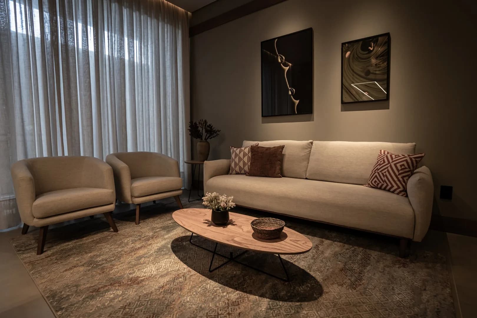 Sala de estar do condomínio com sofá, duas poltronas, mesa de centro oval e cortinas de voil